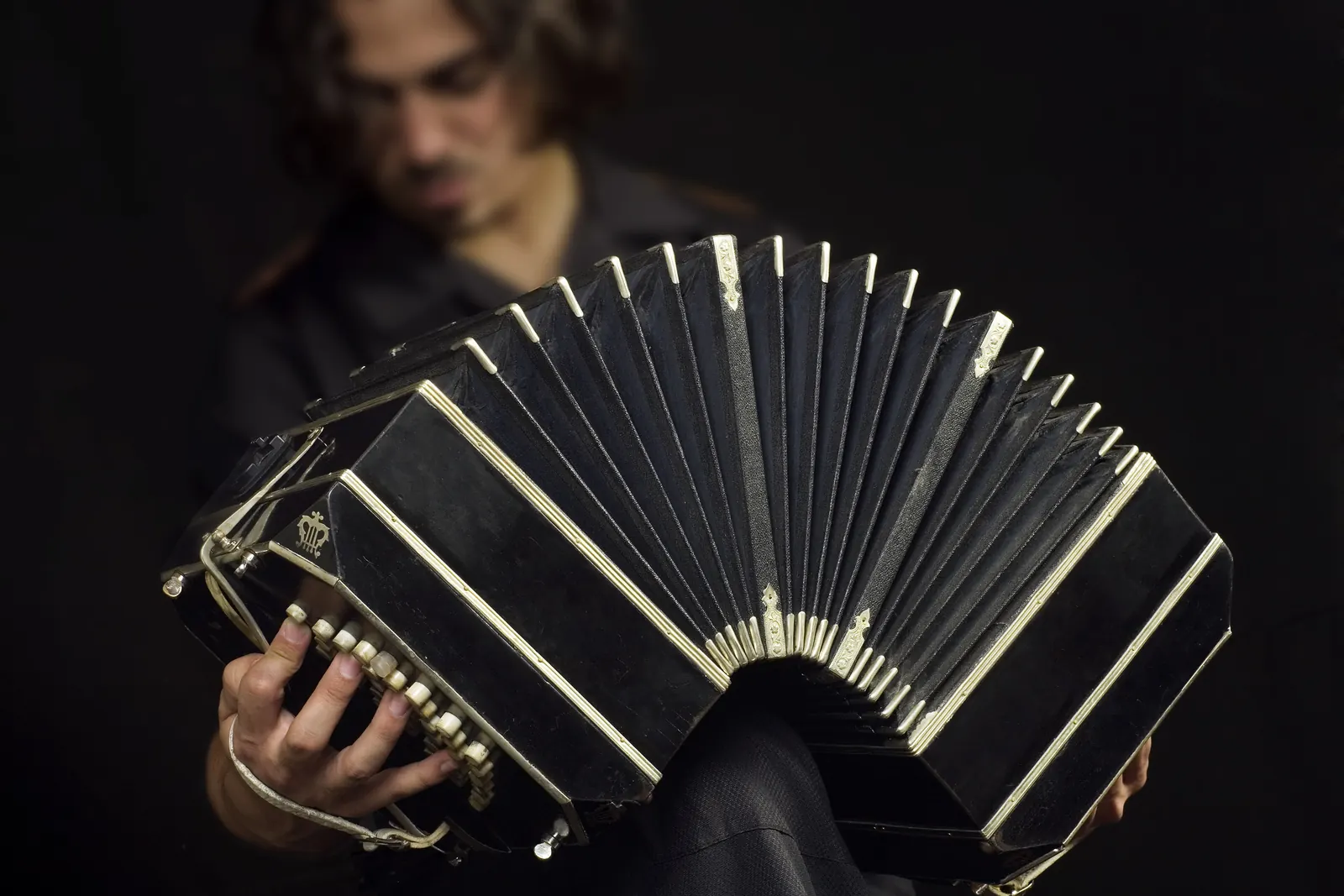 A bandoneón player holding the black instrument with its bellows fully extended, against a dark background