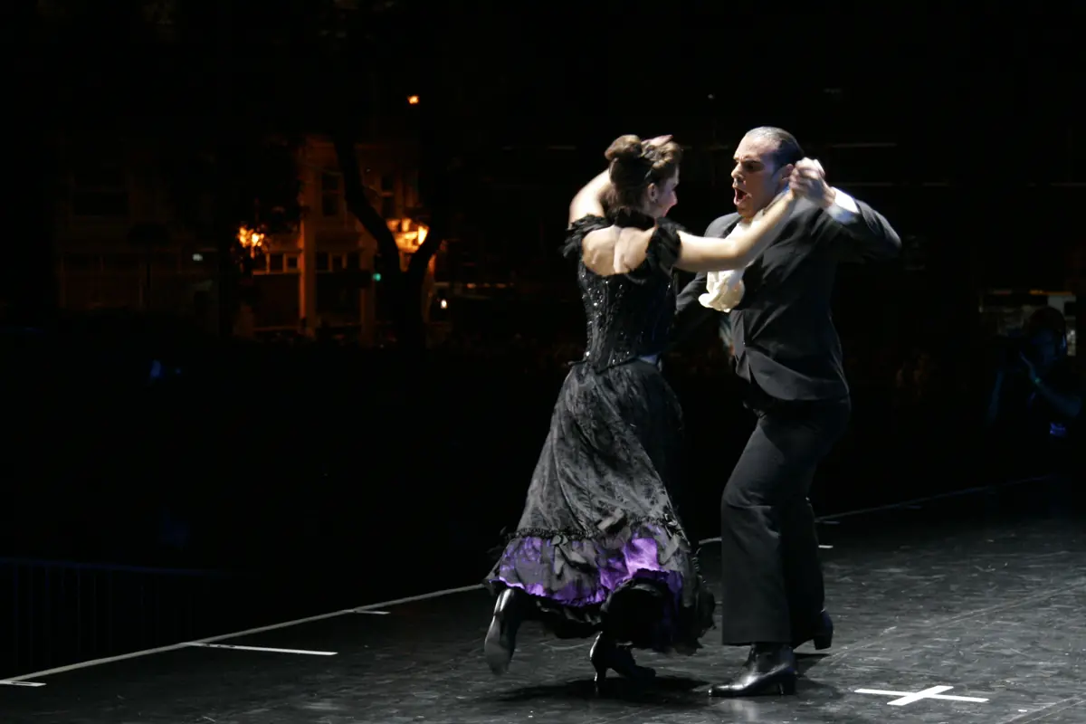A tango couple in black, the woman in a purple-trimmed skirt, dancing on a dark stage lit by a single spotlight