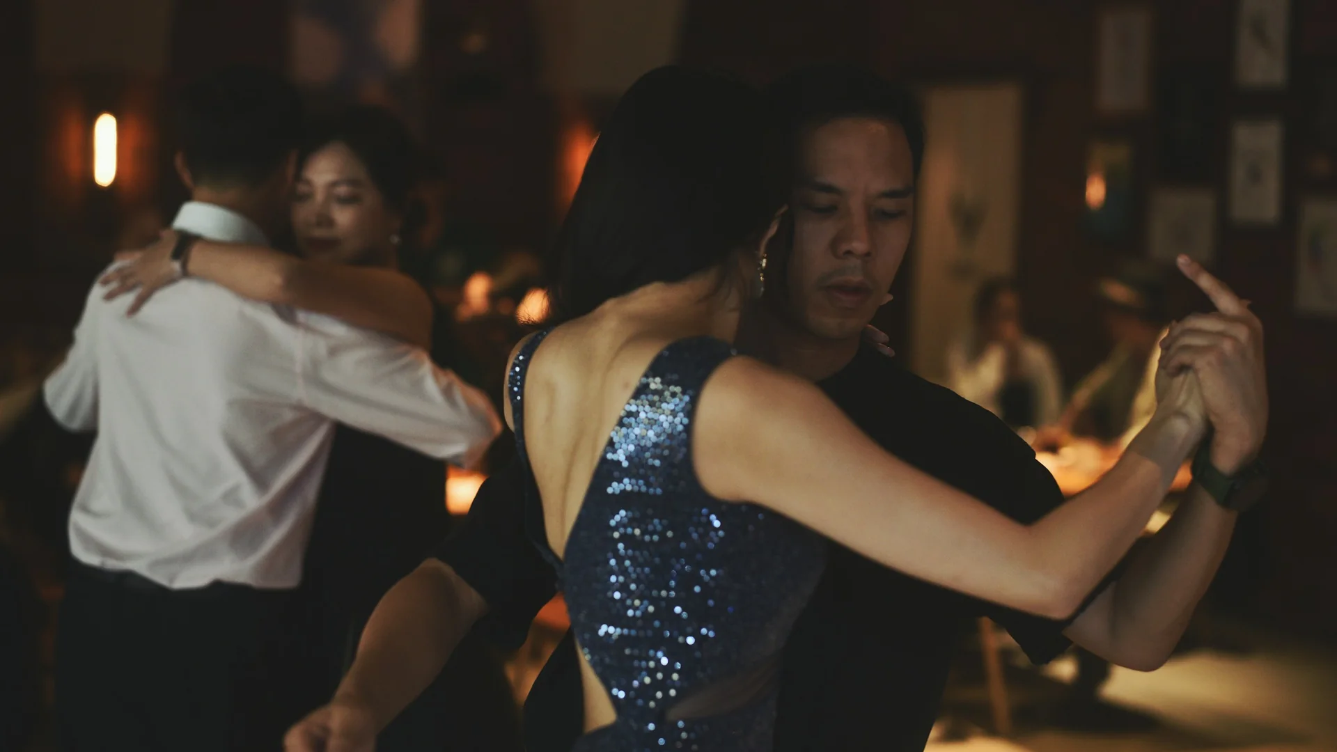 A tango couple in close embrace on a dim dance floor, the woman in a sparkling blue dress, with another couple dancing behind them